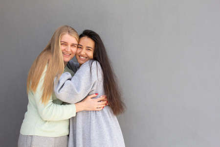 two girls with different color of hair are standing and hugging each other, grey background wall