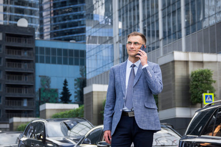 business man stands near the houses and holds the phone in his hands.male portrait on the background of buildings.business man portrait.Man talking on the phone on the street. Man stands near business center.street portrait for advertising.man looking at phone.man with glasses.office worker.の写真素材