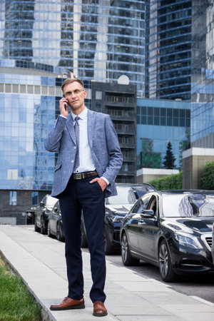 business man stands near the houses and holds the phone in his hands.male portrait on the background of buildings.business man portrait.Man talking on the phone on the street. Man stands near business center.street portrait for advertisingの写真素材