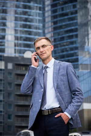 business man stands near the houses and holds the phone in his hands.male portrait on the background of buildings.business man portrait.Man talking on the phone on the street. Man stands near business center.street portrait for advertising.man looking at phone.man with glasses.office worker.の写真素材