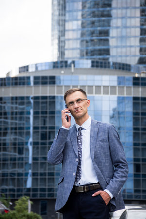 business man stands near the houses and holds the phone in his hands.male portrait on the background of buildings.business man portrait.Man talking on the phone on the street. Man stands near business center.street portrait for advertising.man looking at phone.man with glasses.office worker.の写真素材