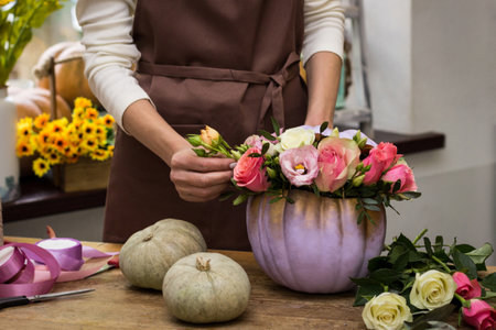 Florist makes an arrangement of pumpkins and flowers.Decoration and floristry training courses.Flower arrangement in pumpkin on a table in store. Flower shop clerk compiling a bouquet on the table.Flowers ribbon scissors pumpkin on the table and in the hands of a florist close-upの写真素材