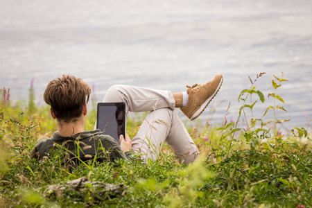 Teenager in nature looking at laptop.A teenager lies in the grass with tablet.The guy near the river lies in the grass.The guy with his back to the frame lies on the river bank with tablet in his handsの写真素材