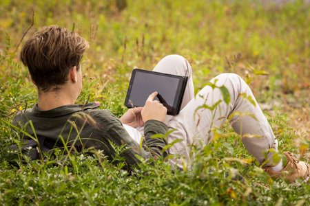 Teenager in nature looking at laptop.A teenager lies in the grass with tablet.The guy near the river lies in the grass.The guy with his back to the frame lies on the river bank with tablet in his handsの写真素材