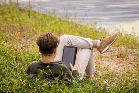 Teenager in nature looking at laptop.A teenager lies in the grass with tablet.The guy near the river lies in the grass.The guy with his back to the frame lies on the river bank with tablet in his handsの写真素材