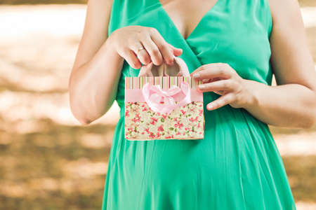 Pregnant woman in green dress pulls out a pink baby booties from a package in the autumn park. Close Up.の写真素材