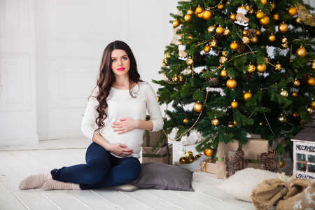 Happy young pregnant woman sitting near the Christmas tree.の写真素材