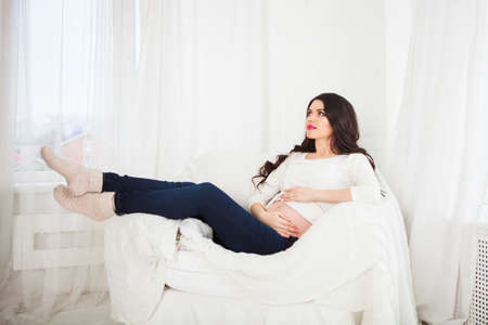 Beautiful pregnant woman sitting at sofa and keeping hand on belly.の写真素材