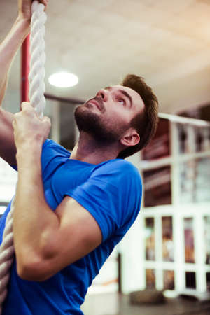 Portrait of young muscular athlete climbing fitness rope in sport club. Male bodybuilder exercising in the gym.の写真素材