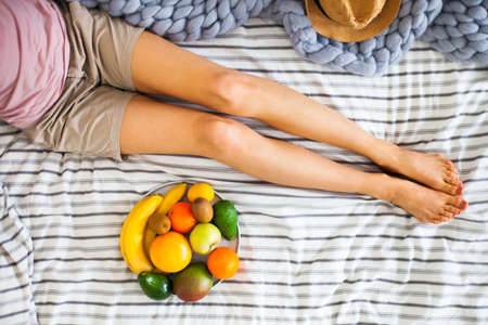 Summer healthy raw vegan clean eating breakfast in bed concept. Young girl wearing pastel colored home clothes taking fruits from tray full of fresh fruit. Top view, square crop, copy spaceの写真素材