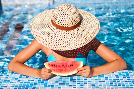 A girl holds half a red watermelon  over a blue pool, relaxing on spa in a tropical hotel, eating...