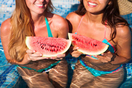Two happy girlfriends holds half a red watermelon  over a blue pool, relaxing on spa in a tropical hotel, eating healthy, fruit diet, summer party styleの写真素材