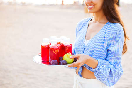 Happy woman holds a dish with a drinks red juice at sunset. Picnic theme on the beach, food and drink conceptの写真素材