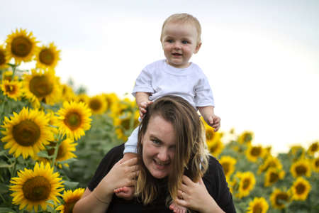 brunette woman young mom with cute baby blonde son on a field of sunflowers.