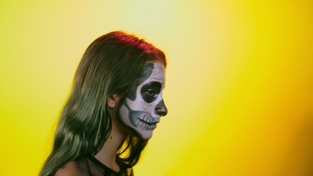 Halloween, girl with make-up skeleton on half face, dressed as witch, posing on bright yellow...