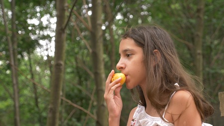 Cute little girl brunette, eating fruit and talking. Nature background.の写真素材