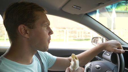the man at the wheel of the car, eats fast food, while the car is moving. in a summer sunny day.の写真素材