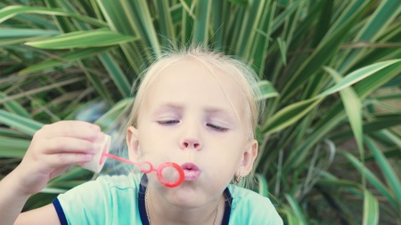 little blond girl blowing soap bubbles against a tropical park backgroundの写真素材