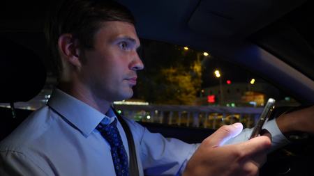 Texting with a smart phone in car. a young businessman is driving a car around the night city and...