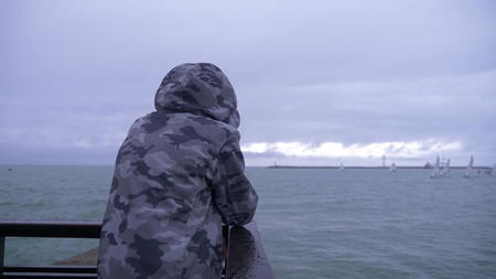 someone in a hooded jacket is standing on the pier and watching the sailboats sailing to the sea.