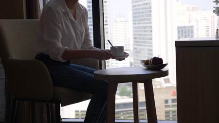 Beautiful woman drinking coffee by the window overlooking the city, skyscrapers.の写真素材
