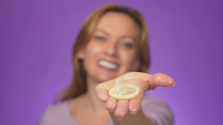 Young beautiful woman, blurred in the background, smiling. in focus is her hand holding an open condom, the concept of safe sex. Protection against AIDS and birth control, color backgroundの写真素材