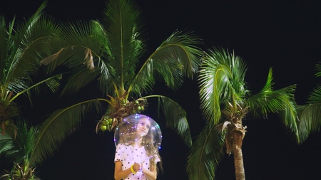 happy teen girl holding big airy, glowing balloon in her hands against the background of palm trees....