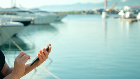 female hand holds smartphone on blurred background of port with yachtsの写真素材