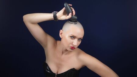 a cool sexy bald woman in a leather corset shaves her head with an electric razor against a dark background. copy space. adventures of strange people.の写真素材