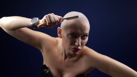 A cool sexy bald woman in a leather corset shaves her head with a dangerous razor on a dark background. Copy space. adventures of strange people.の写真素材