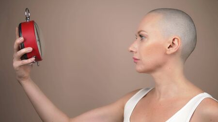 Portrait of shocked beautiful bald woman with an alarm clock on a beige background.の写真素材