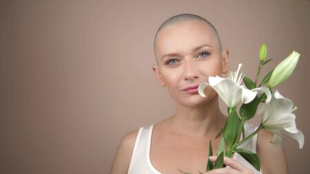 beautiful smiling bald girl sniffs a white lily. beige background, copy space