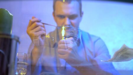 a man is preparing a drug in a spoon, holding it over the flame of a lighter. view through a glass...