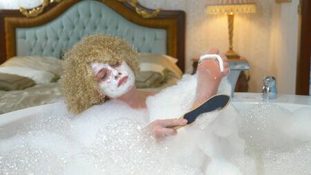 cheerful blonde woman does pedicure while taking a bath with foam