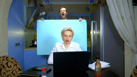 A woman works remotely from home in the kitchen, her husband is holding a blue background. humorの写真素材