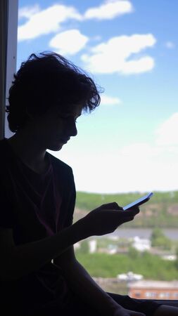 vertical. silhouette. The boy uses a phone, by the window against the blue skyの写真素材