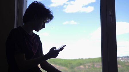 silhouette. The boy uses a phone, by the window against the blue sky and hillsの写真素材