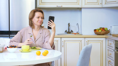 woman eating while sitting in the kitchen at the table and using her smartphoneの写真素材