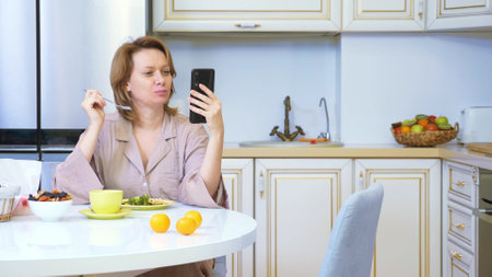 woman eating while sitting in the kitchen at the table and using her smartphoneの写真素材