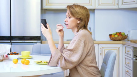 woman eating while sitting in the kitchen at the table and using her smartphoneの写真素材