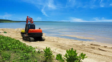 Excavator or digger moving beach sand after erosion by the tides on a tropical beach with calm ocean...