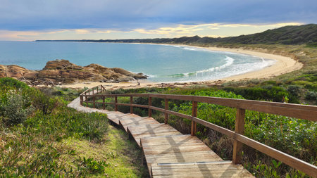 At Cape Conran Victoria Australia a wooden boardwalk winds down to a tranquil seaside cove, revealing a curving sandy beach, clear blue water, rocky outcrops, and lush coastal vegetation under a cloudy sky.の写真素材