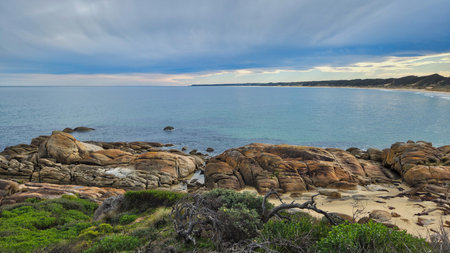 At Cape Conran Victoria Australia view of a tranquil seaside cove, revealing a curving sandy beach, clear blue water, rocky outcrops, and lush coastal vegetation under a cloudy sky.の写真素材