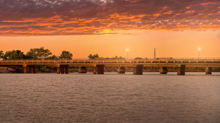Norman River, warm sunset fills the sky over a long bridge spanning a the Norman river. Lamps glow along the roadway as trees outline the banks, creating a tranquil, scenic moment.の写真素材