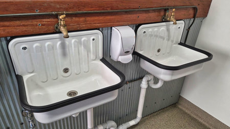 A row of white sinks with brass taps and exposed plumbing along a wooden back splash in a functional utility room. Clean, practical design emphasizes sanitation and maintenance in a shared space.の写真素材