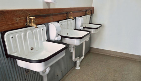 A row of white sinks with brass taps and exposed plumbing along a wooden back splash in a functional utility room. Clean, practical design emphasizes sanitation and maintenance in a shared space.の写真素材