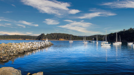 Quarantine Bay Eden New South Wales a tranquil harbor scene features moored sailboats, calm reflective water, a blue cloudy sky over distant trees. Ideal for travel, nature, or nautical themes and peaceful ambiance.の写真素材