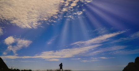 A lone hiker stands on a grassy hill as sun rays spill through the clouds, creating a dramatic, expansive sky. Ideal for outdoor, travel, and nature themes.の写真素材