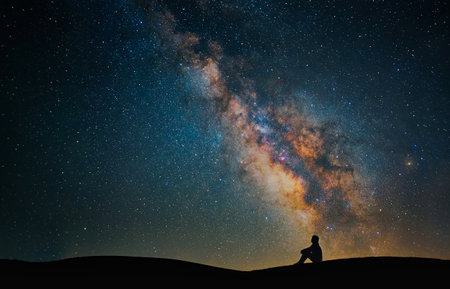 A lone silhouette sits on a dune beneath a star-filled night sky, with the Milky Way stretching overhead. The image conveys wonder, solitude.の写真素材
