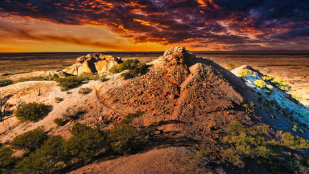 A dramatic desert landscape at sunset features eroded mounds, sparse shrubs, and a fiery sky....